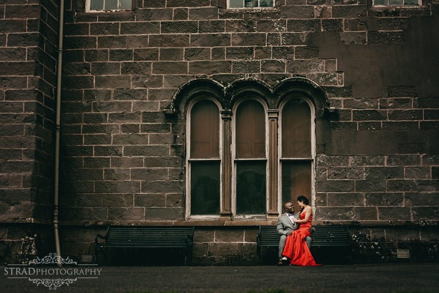 Bride in the red dress sitting on the grooms lap with a big wall behind