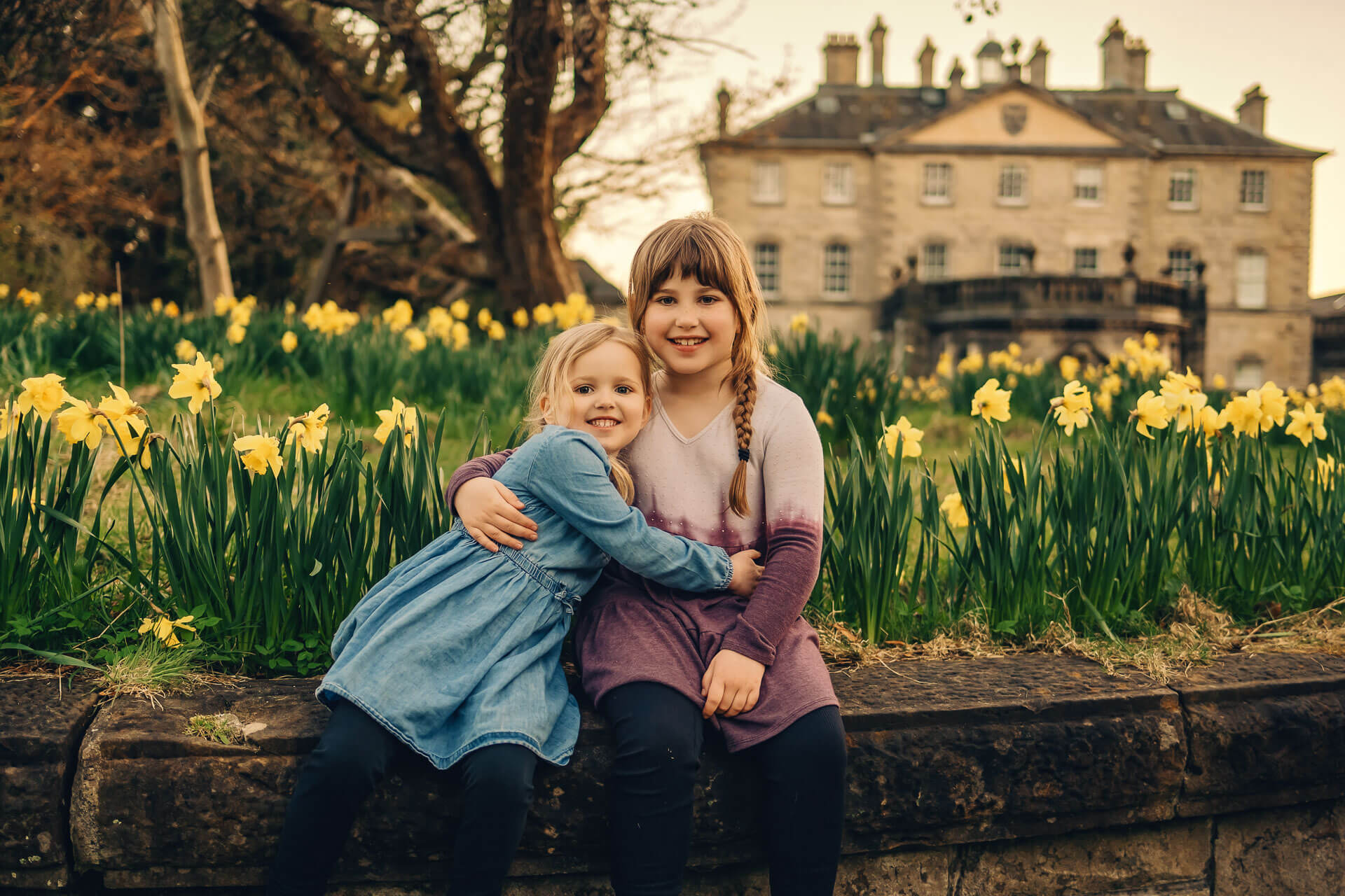 Two sisters hugging beside daffodils with Pollok House in Glasgow behind them