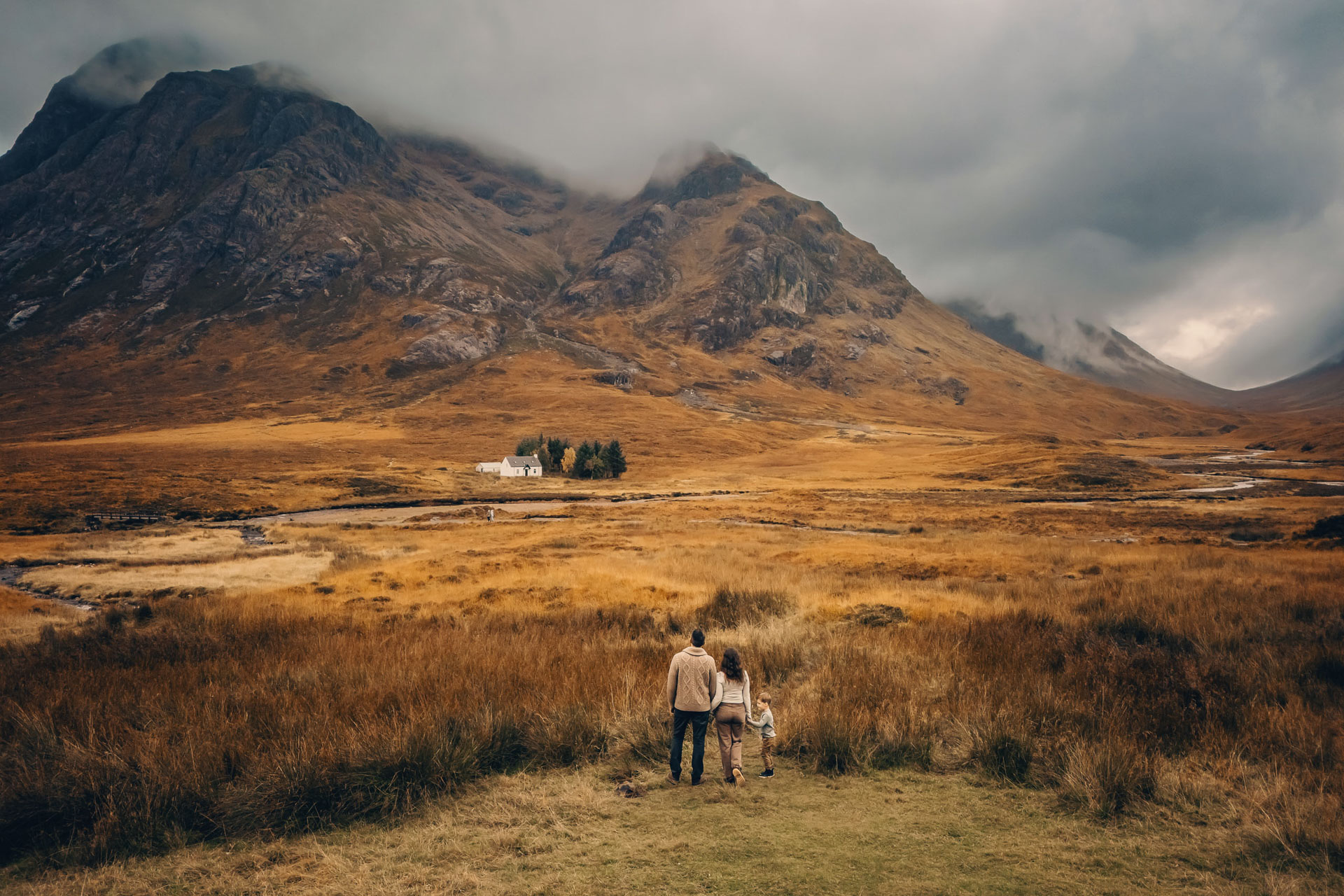 a drone photo of a family of 3 walking in a field with mountains in the background in Glencoe