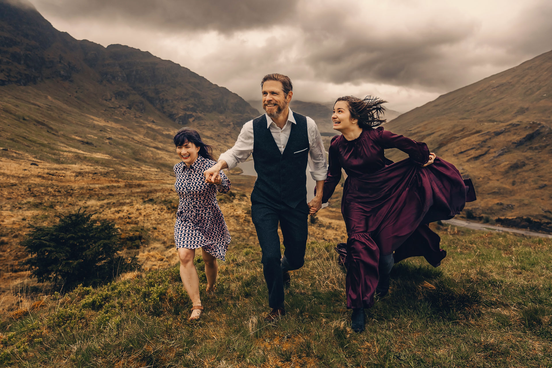 a mother and father with teenage daughter in a long dress are running on a hill with mountains in the background at the rest and Be Thankful viewpoint.