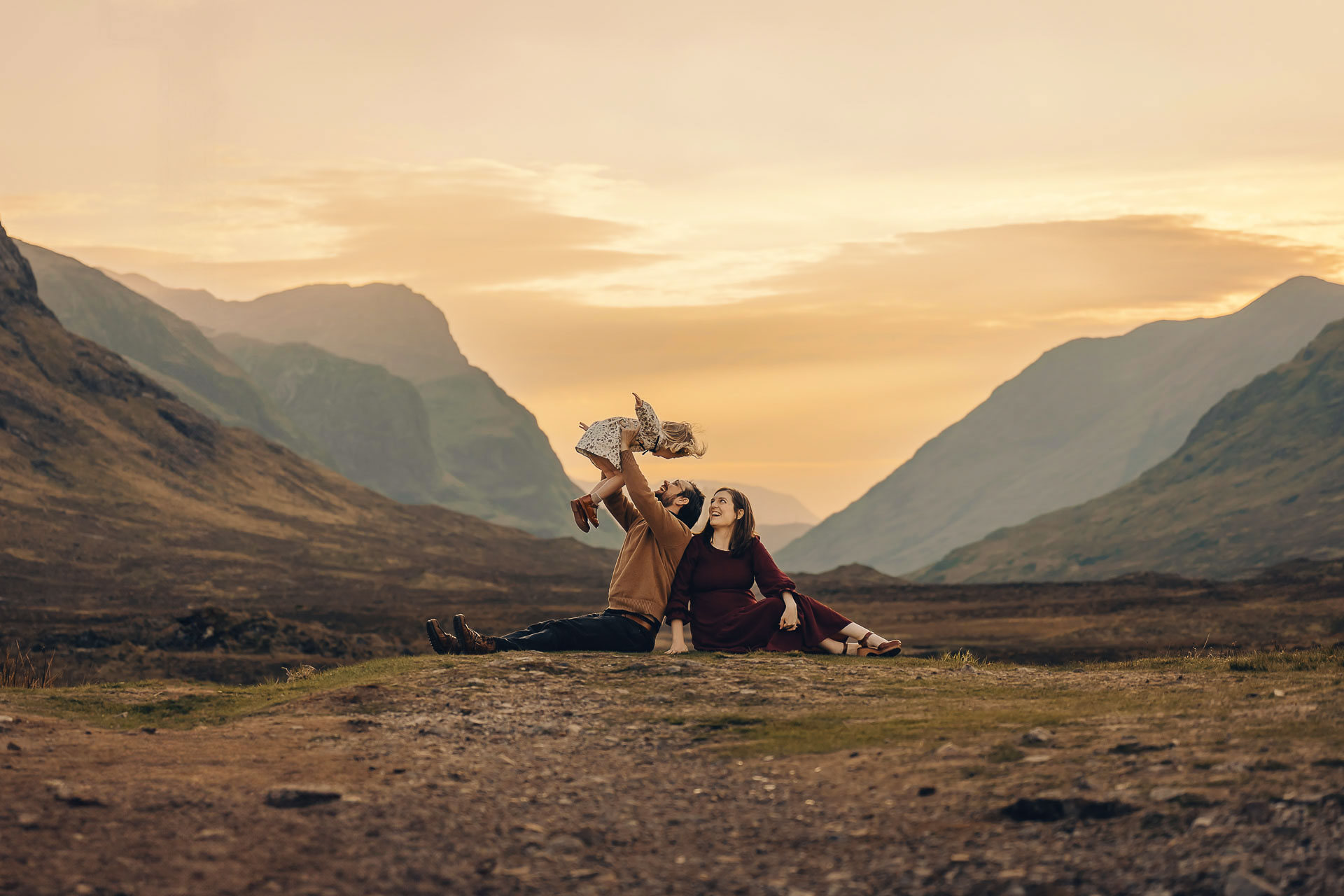 a man and woman sitting on a hill lifting child in the air