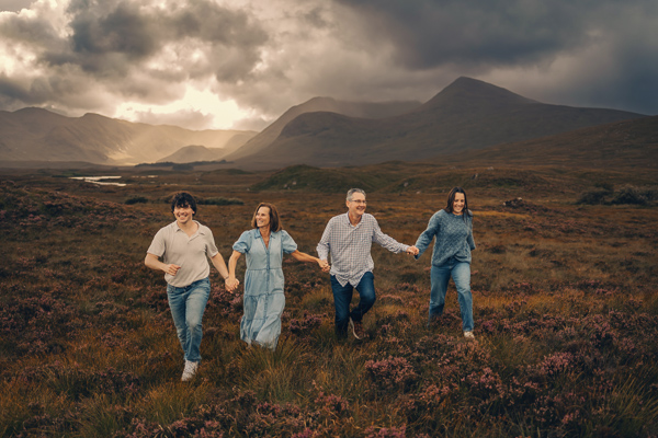 A family of four is running together through the grass in Glencoe with dramatic sky and mountains behind them.