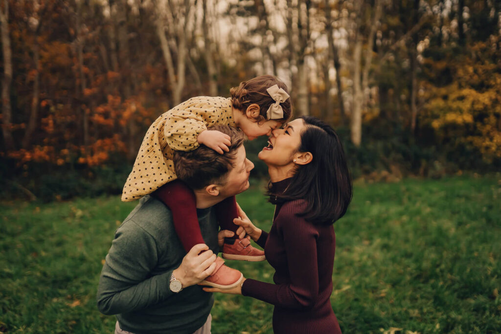 Dad holding daughter on his shoulder while she kisses her mum in the park in Glasgow.