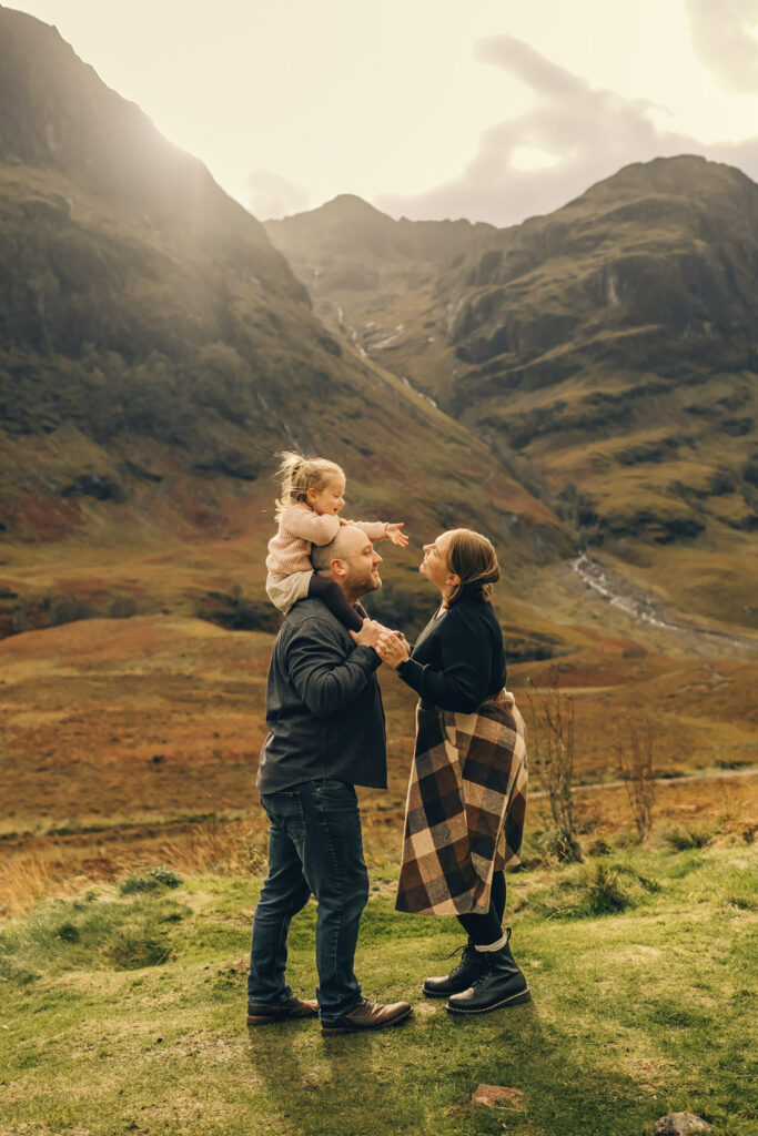 Dad is holding his daughter on his shoulder while daughter is talking to her mum with Three Sisters of Glencoe behind them.