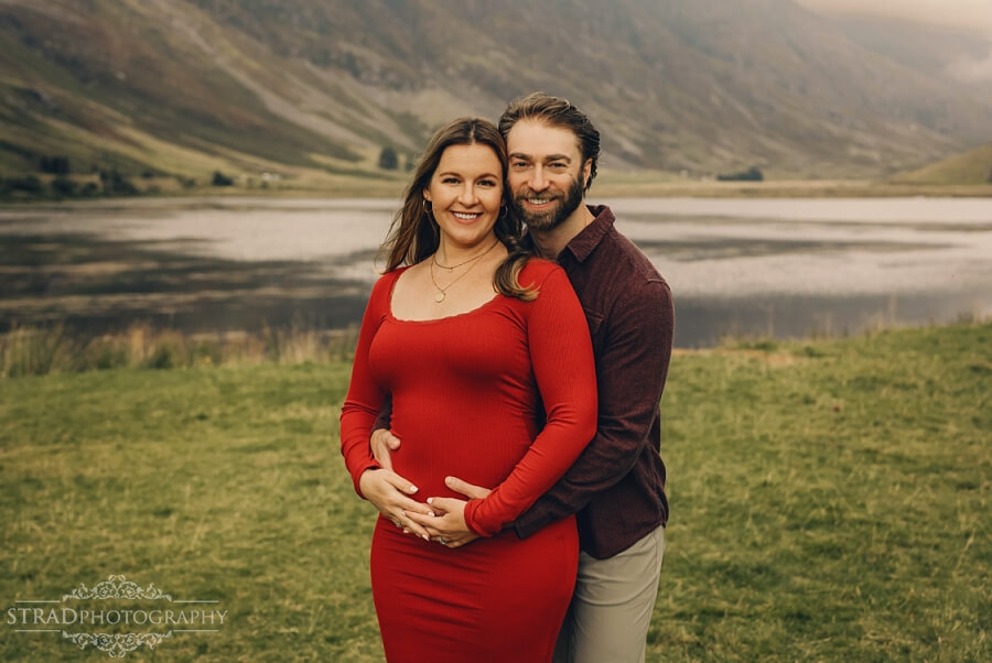Man cuddling his pregnant wife by scenic landscape near Loch Achtriochtan.