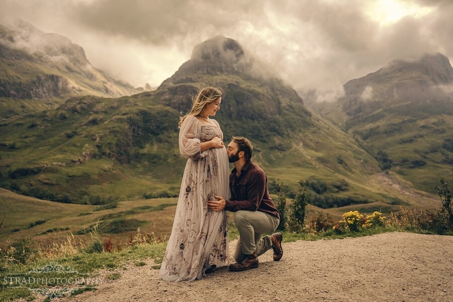 Father kisses his pregnant wife belly in scenic mountain landscape in Glencoe.