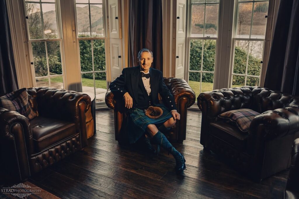 Man in kilt seated, elegant setting.